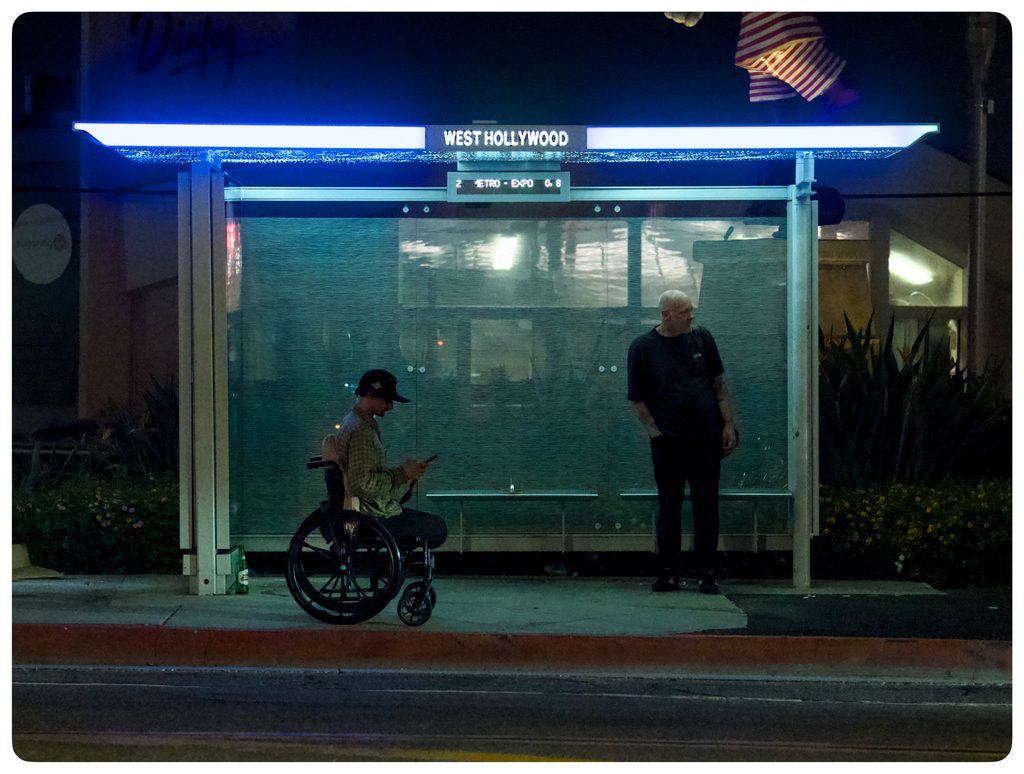 Night at a glowing West Hollywood bus shelter: a man in a wheelchair checks his phone while another man stands waiting